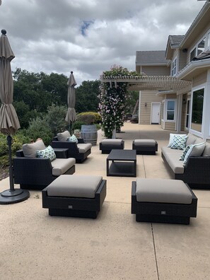 Un patio exterior con mobiliario de mimbre, incluyendo sofás y una mesa de centro, y una pérgola adornada con flores.