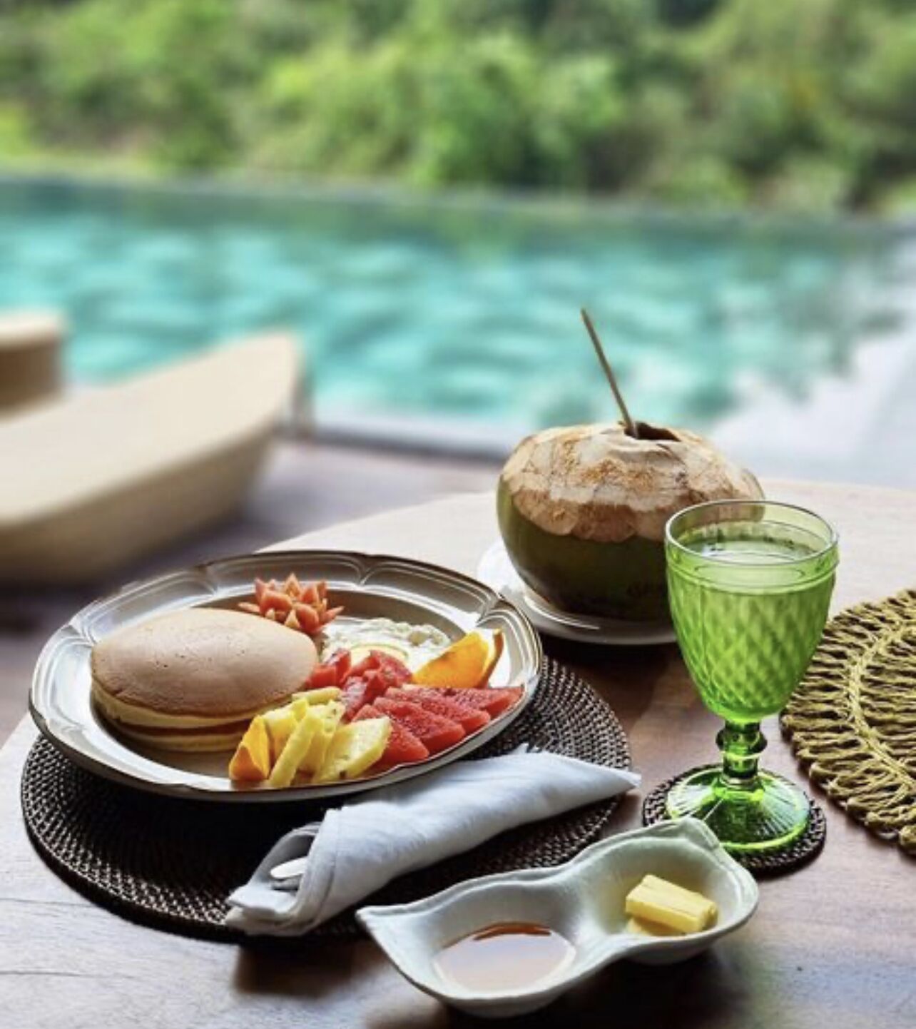 A breakfast setting with pancakes, fruit, a coconut drink, and a butter dish by a pool.