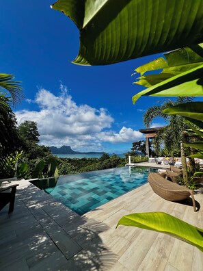 Ein Pool ohne Ende mit klarem, blauem Wasser, umgeben von tropischer Vegetation und Blick auf Berge und das Meer.