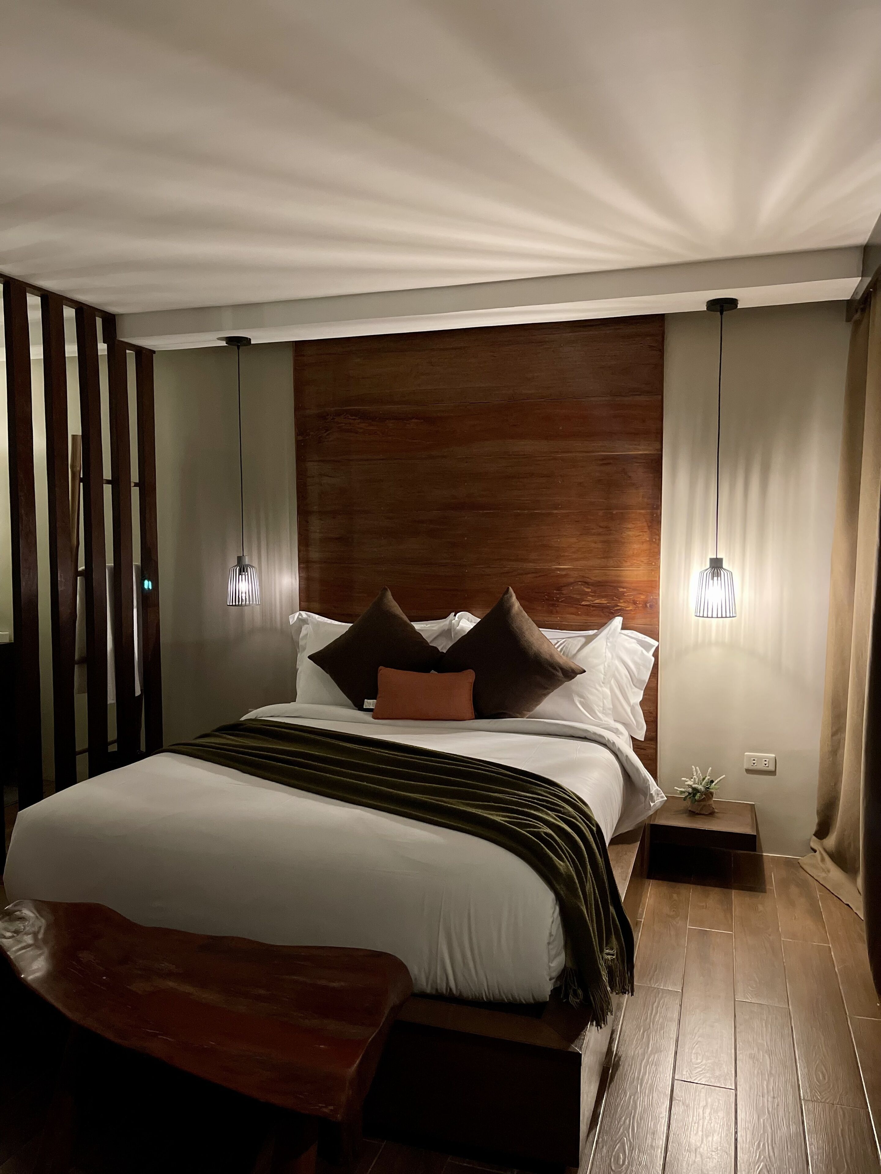 A modern bedroom with a wooden headboard, a large bed with white linens, two bedside lamps, and a wooden bench at the foot of the bed.