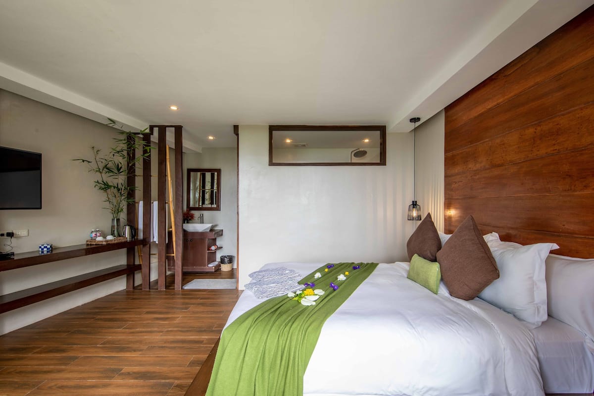 Habitación de hotel con piso de madera, una cama con ropa blanca y almohadones decorativos, un plaid verde, cabecera de madera, televisor montado y repisa de madera con varios objetos.