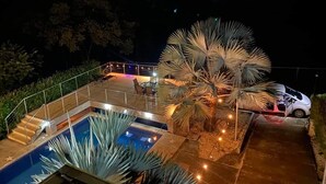 Vista nocturna de un área de piscina con una palmera, un coche y una terraza con sillas.