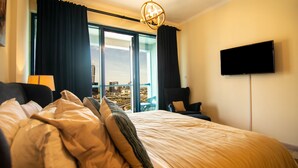 Habitación de hotel con una cama grande, un televisor de pantalla plana, vista a un paisaje urbano a través de un ventanal amplio y un candelabro.