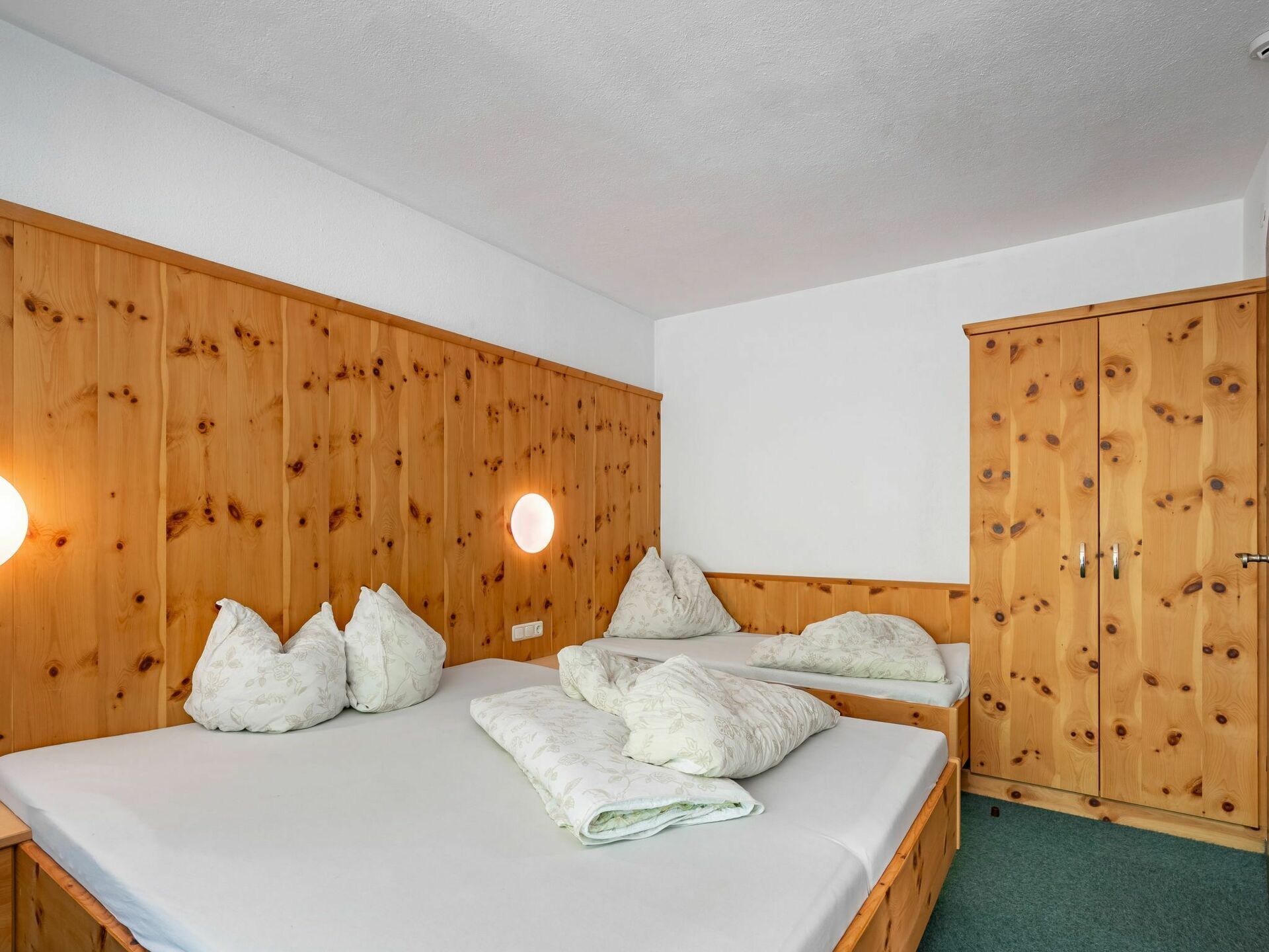 A room with a wooden bunk bed, two bedside tables, a wardrobe, and a wall-mounted light fixture.