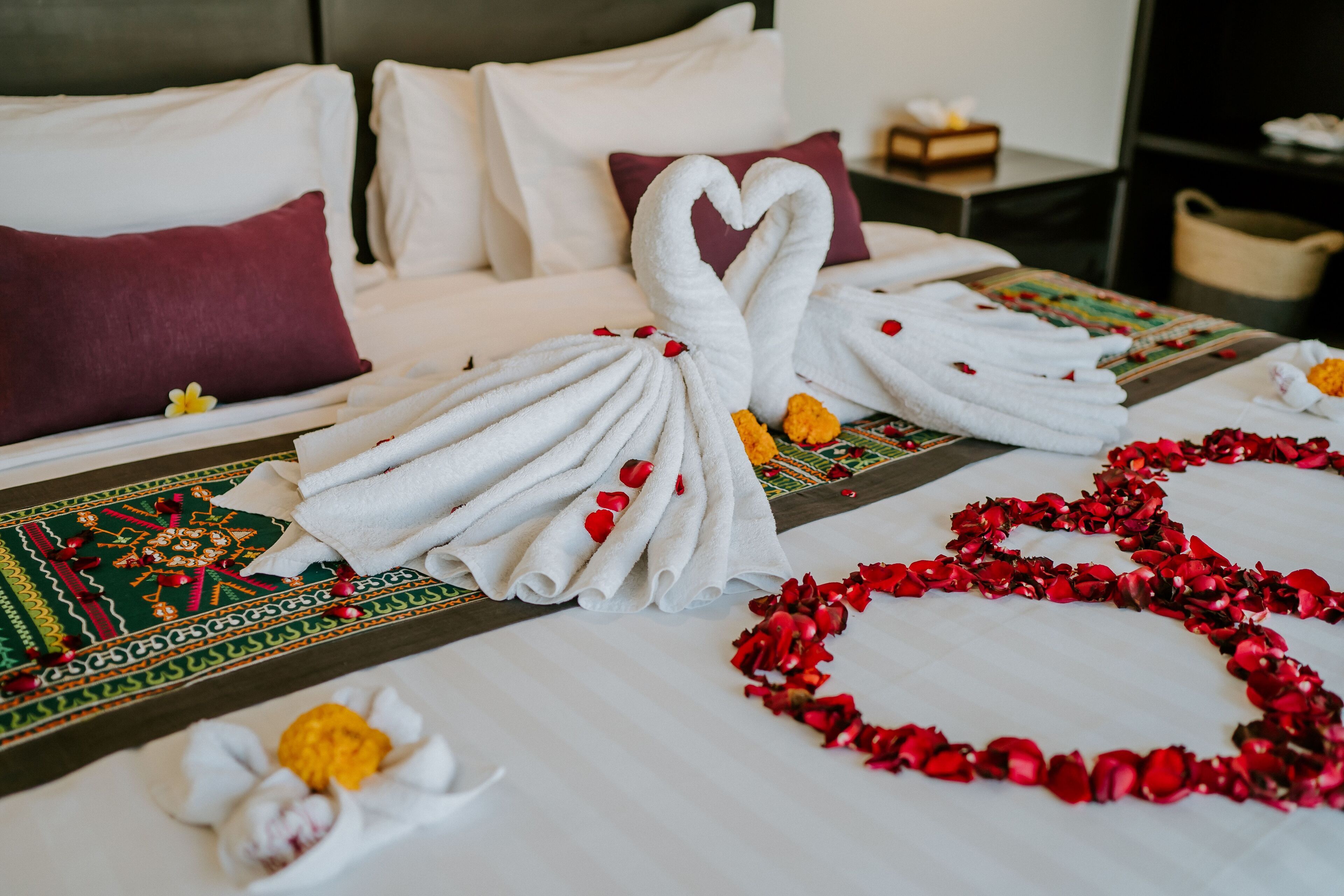 A neatly made bed with white towels arranged in a heart shape, red rose petals, and a decorative pillow.