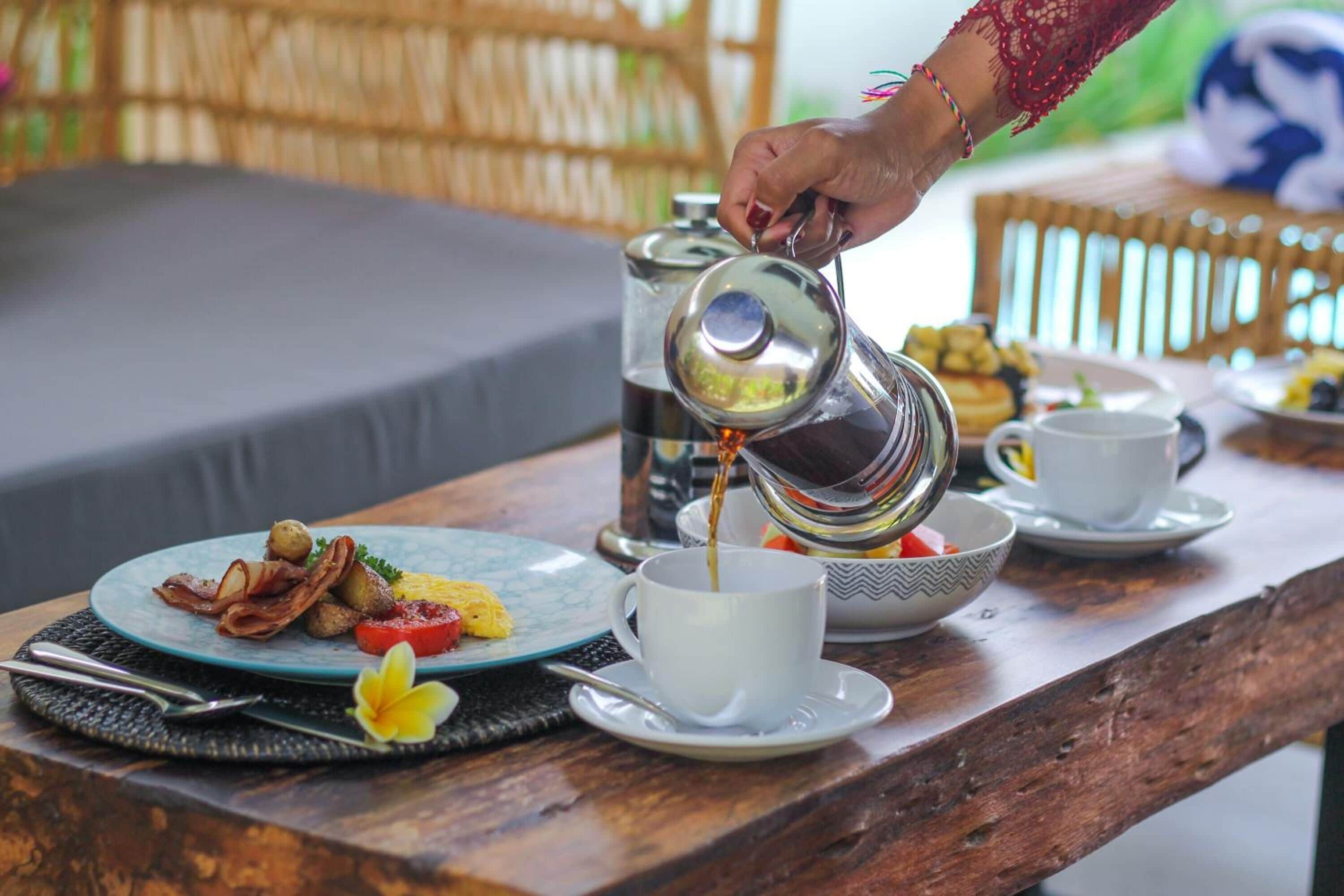 A breakfast setup with a coffee pot pouring into a cup, plates with food, and a wicker chair in the background.