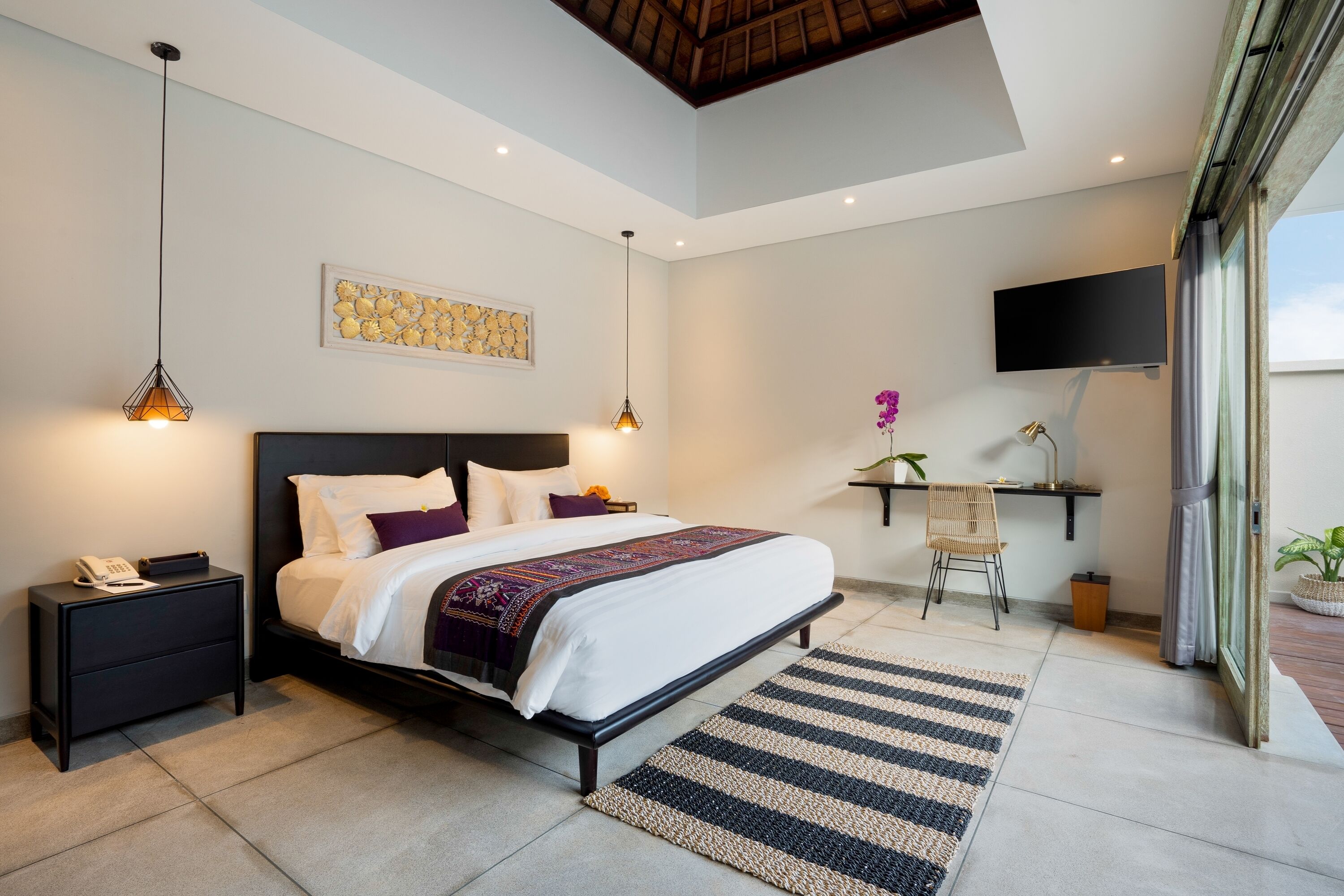 A modern bedroom with a large bed, bedside tables, a desk with a chair, a flat-screen TV, and a large window.