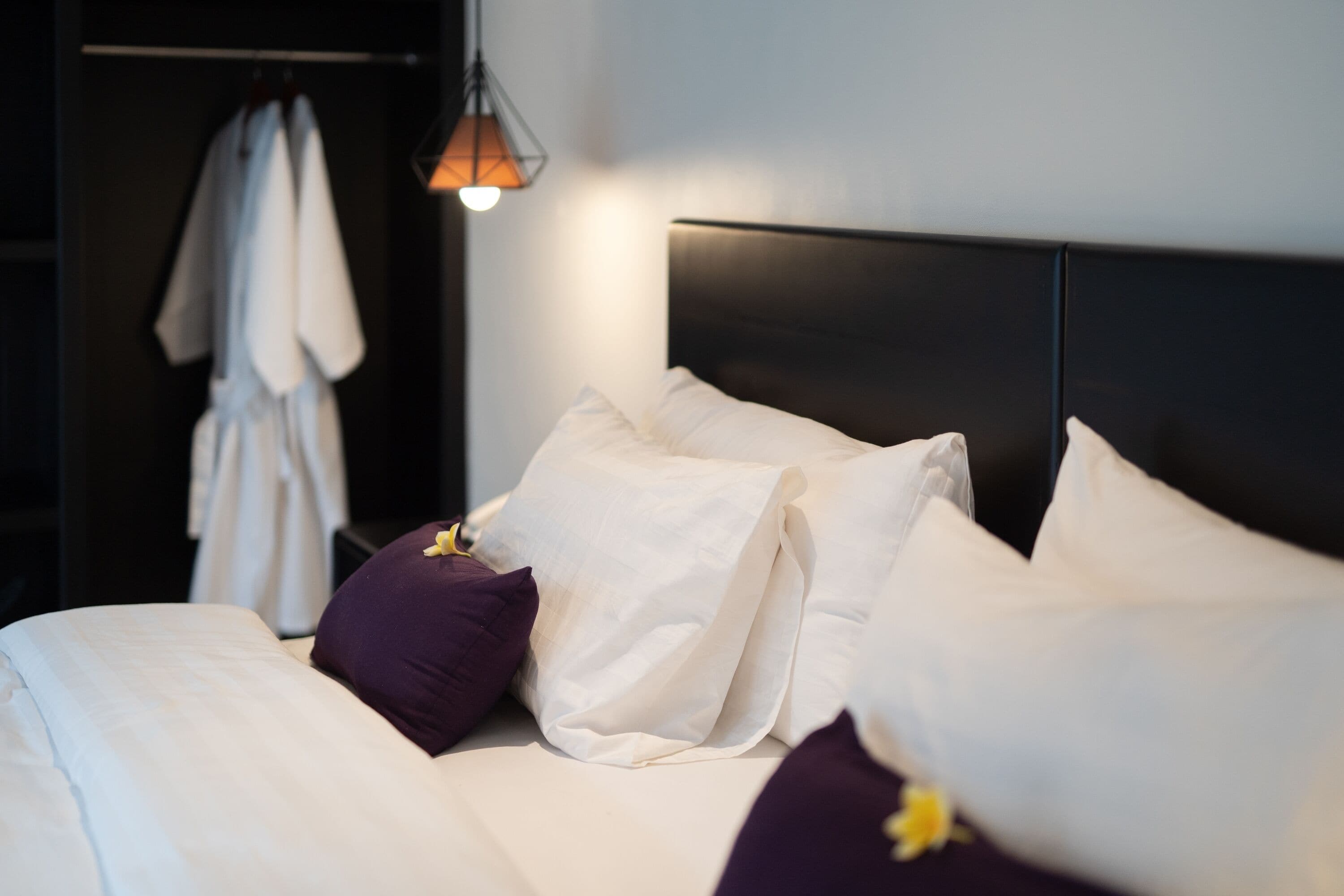 A neatly made bed with white and purple pillows, a dark headboard, and a hanging light fixture.