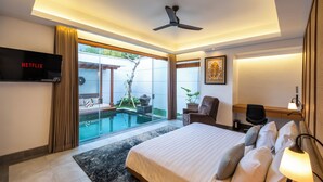 A modern hotel room with a large bed, a flat-screen TV, a ceiling fan, a desk with a chair, a small table, and a large window with a view of a swimming pool and outdoor seating.