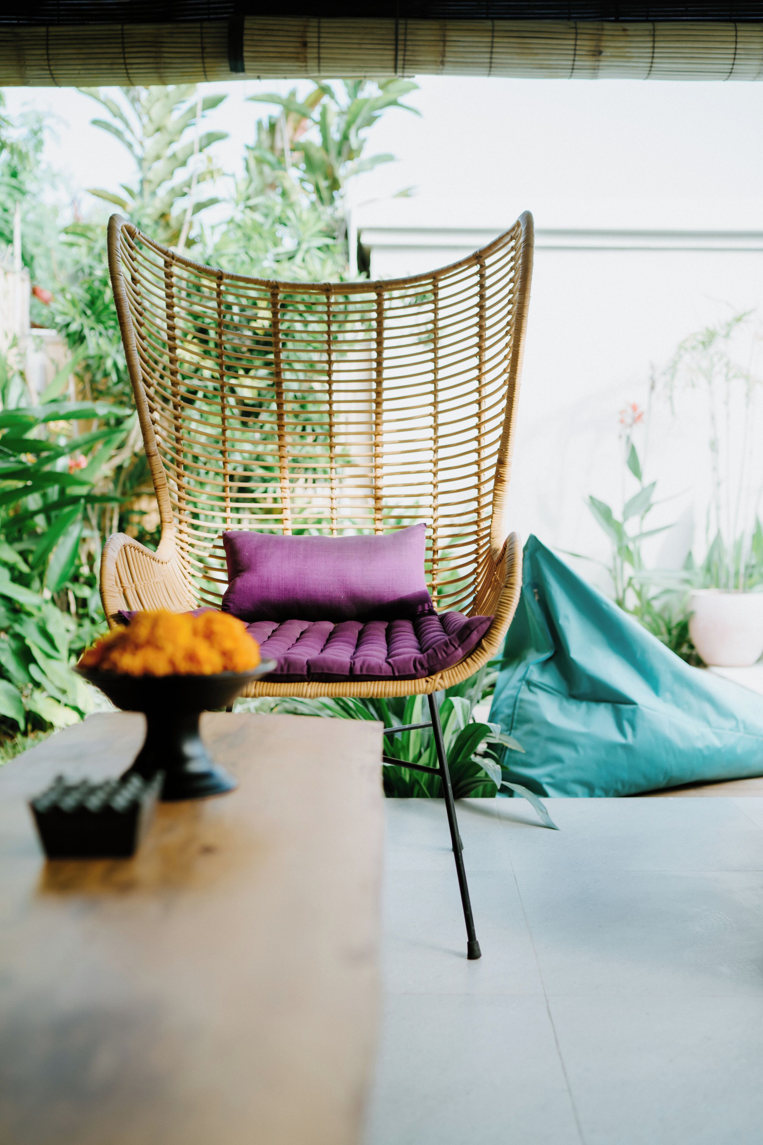 A wicker chair with a purple cushion, a teal bean bag, and a bowl of fruit on a table.