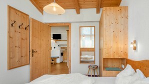 Een slaapkamer met houten wanden en plafond, een bed met wit beddengoed, een klein krukje en een raam met uitzicht op een keuken en een televisie.