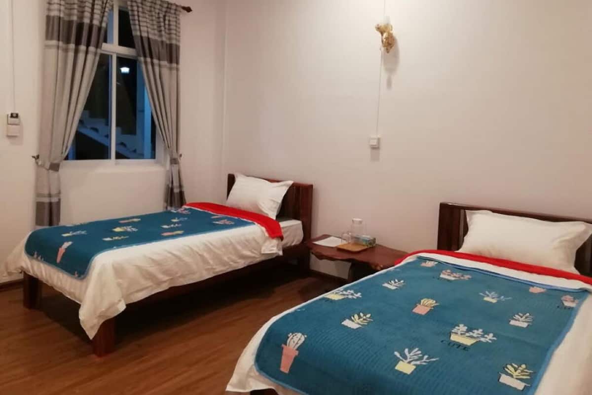 Une chambre d’hôtel avec deux lits simples, une fenêtre avec des rideaux, une table de chevet avec une lampe et un luminaire fixé au mur.