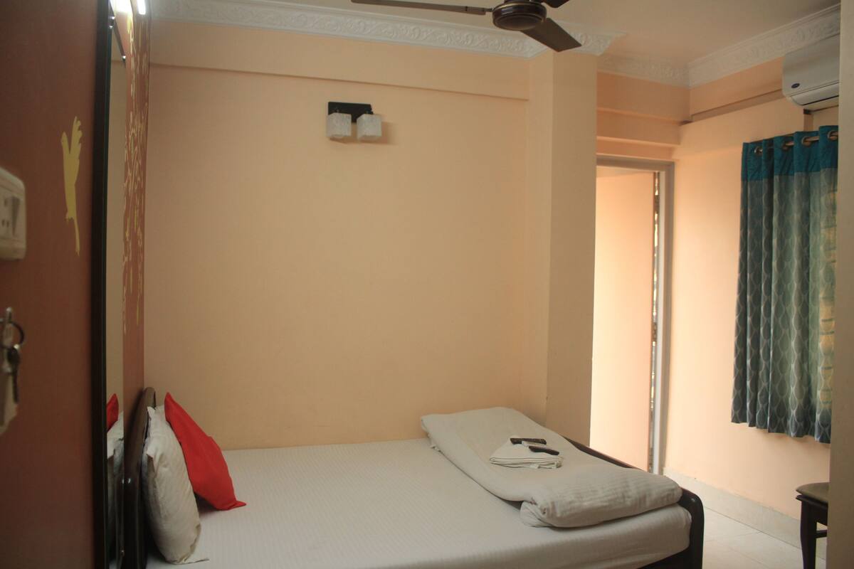 A single bed with a white mattress and pillows, a ceiling fan, a wall-mounted light fixture, and a window with curtains.