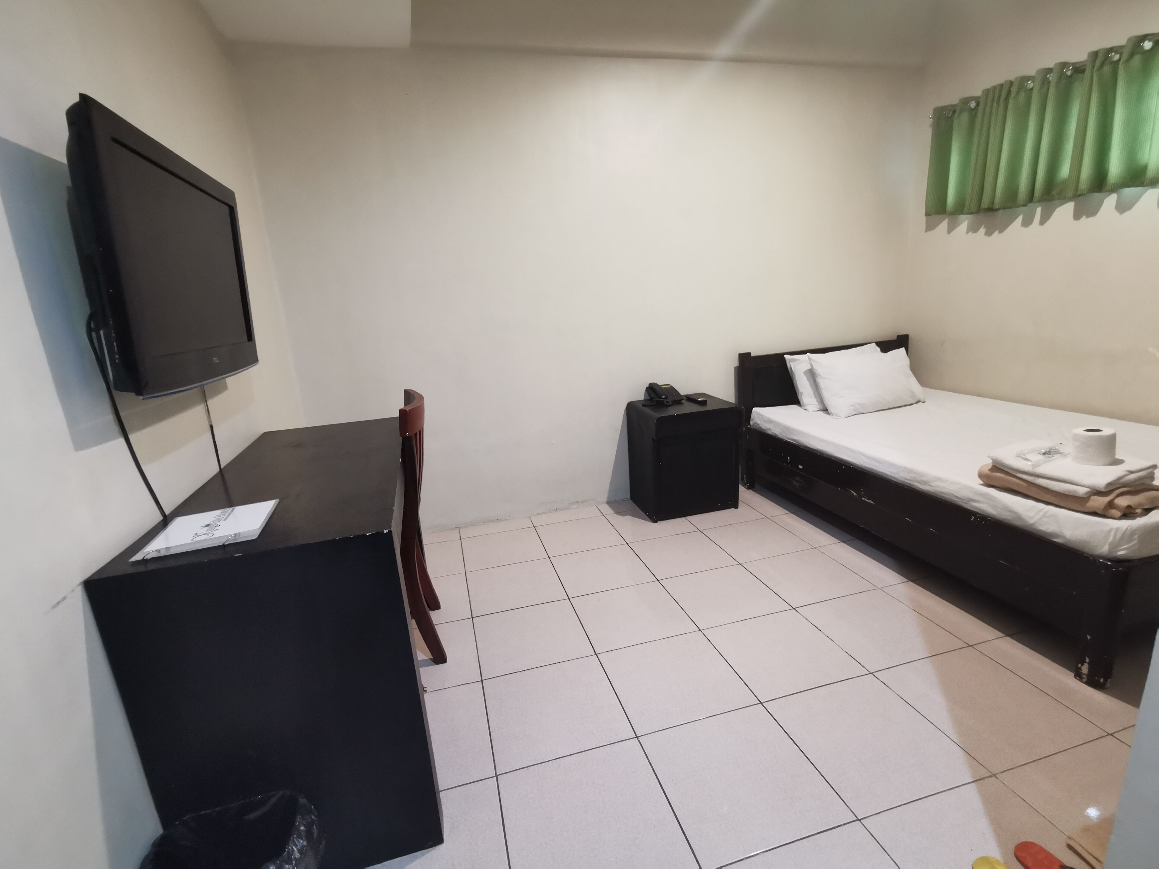 A small, clean room with a bed, a desk, a chair, a TV, and a green curtain.