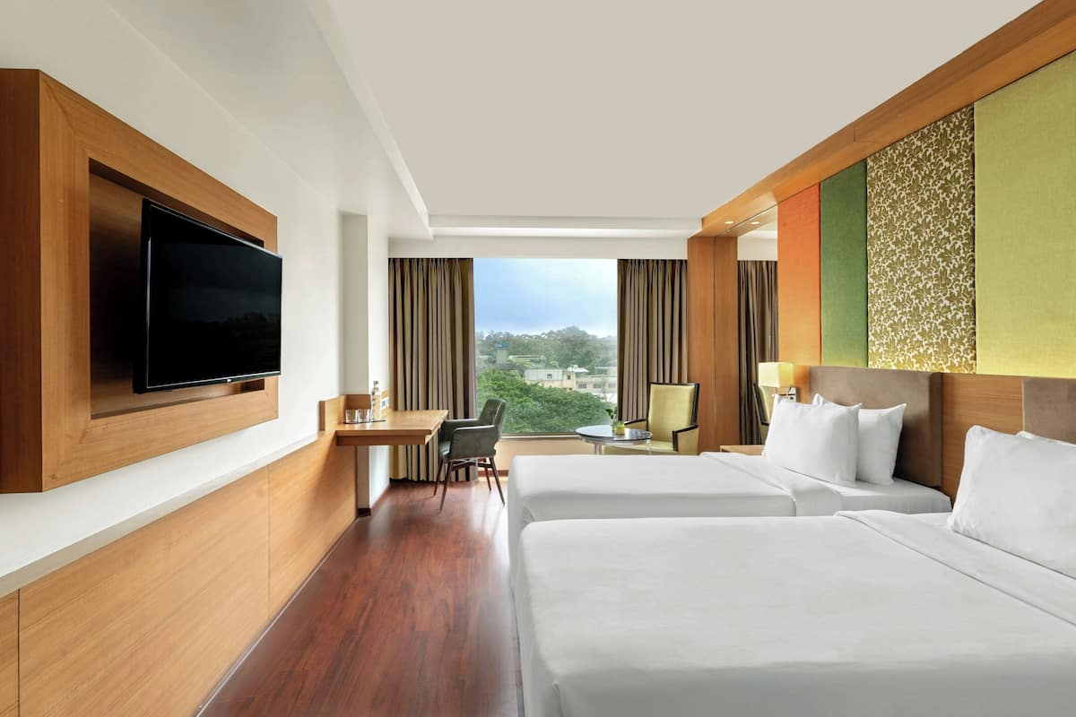 A hotel room with two beds, a desk, a chair, a TV, and a view of the city.