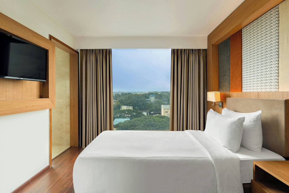 A hotel room with a large bed, a TV mounted on the wall, and a view of the cityscape through the window.