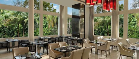 A spacious dining area with multiple tables and chairs, large windows offering a view of lush greenery, and modern red pendant lighting.