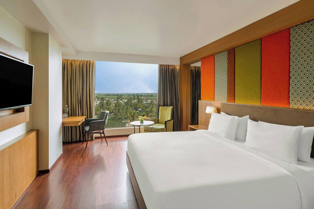 A hotel room with a large bed, a television, a desk with a chair, a small table with a chair, and a view of the landscape.