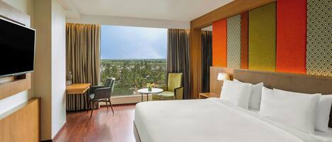 A hotel room with a large bed, a television, a desk with a chair, a small table with a chair, and a view of the landscape.