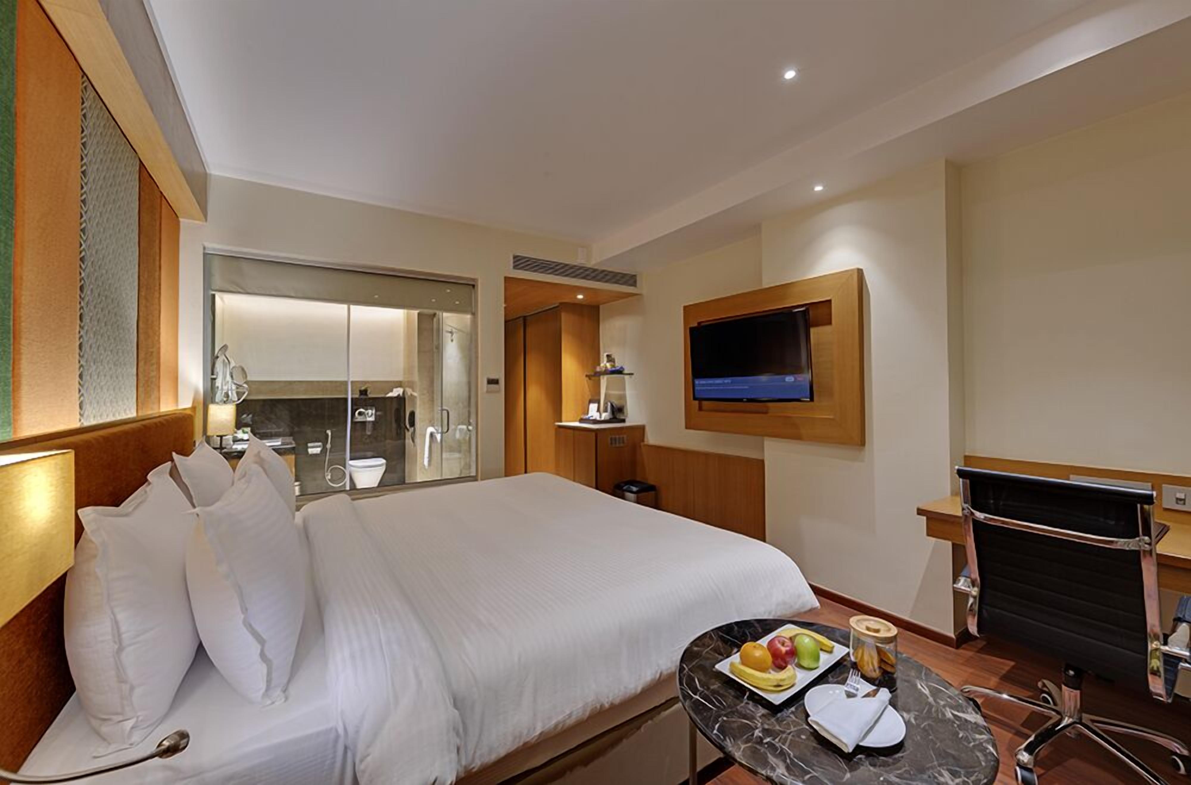 Habitación de hotel con una cama grande, un televisor montado en la pared, una pequeña mesa con fruta y un tarro, y un escritorio con una silla.
