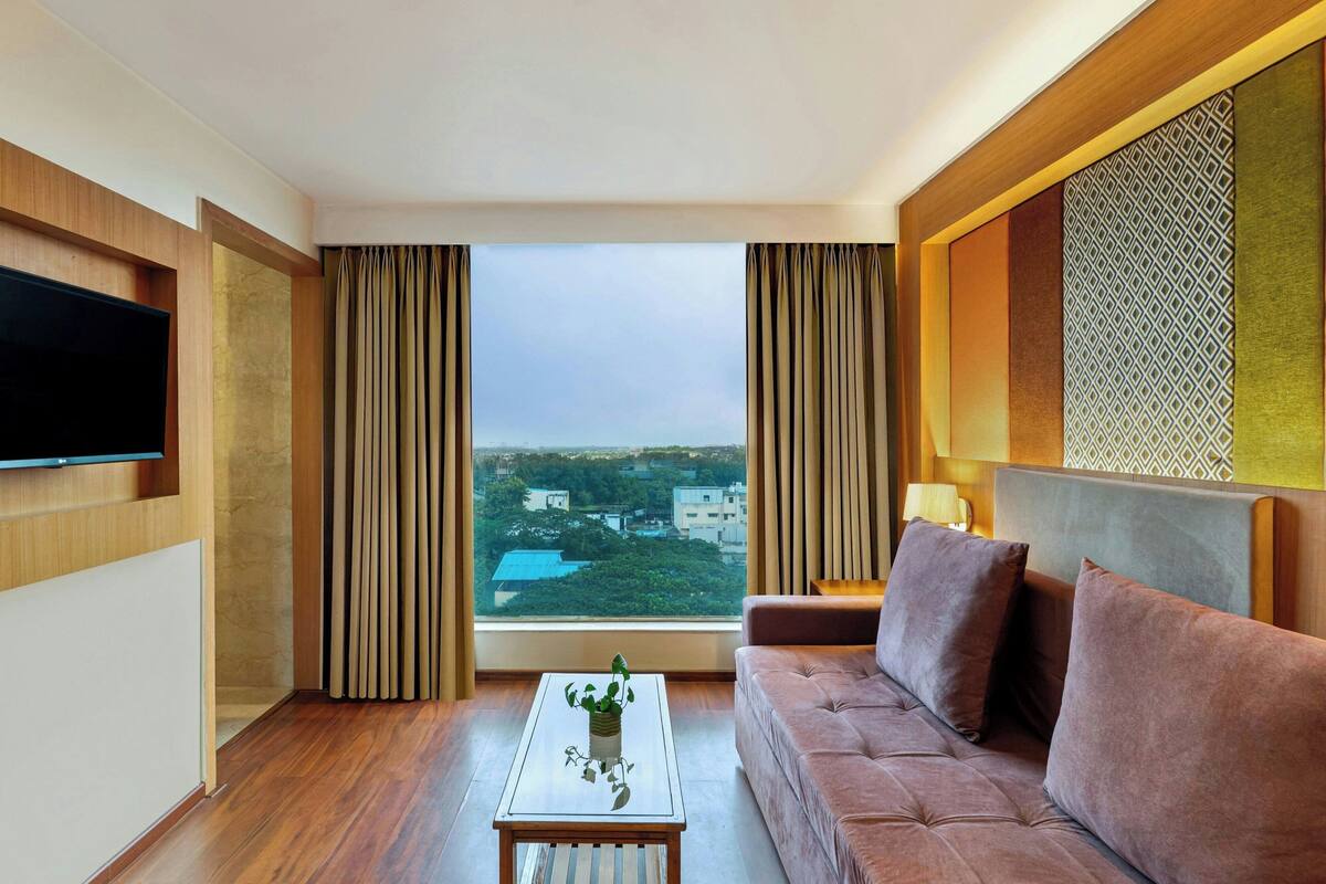 A modern hotel room with a large window, a flat-screen TV, a sofa, and a coffee table.