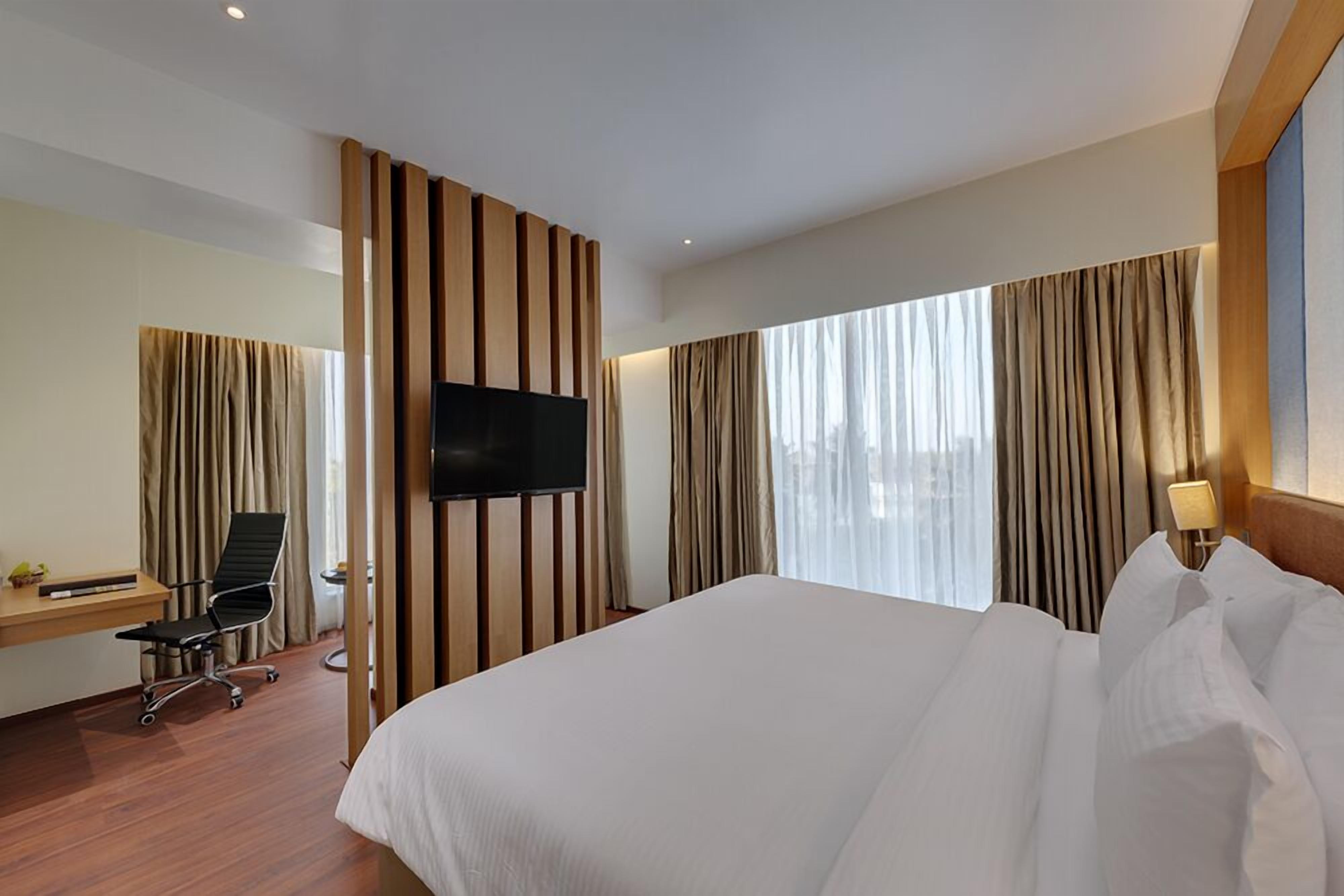 Quarto de hotel com uma cama grande, uma escrivaninha com cadeira, televisão de tela plana na parede e uma janela com cortinas.