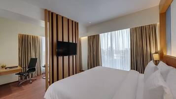 Quarto de hotel com uma cama grande, uma escrivaninha com cadeira, televisão de tela plana na parede e uma janela com cortinas.