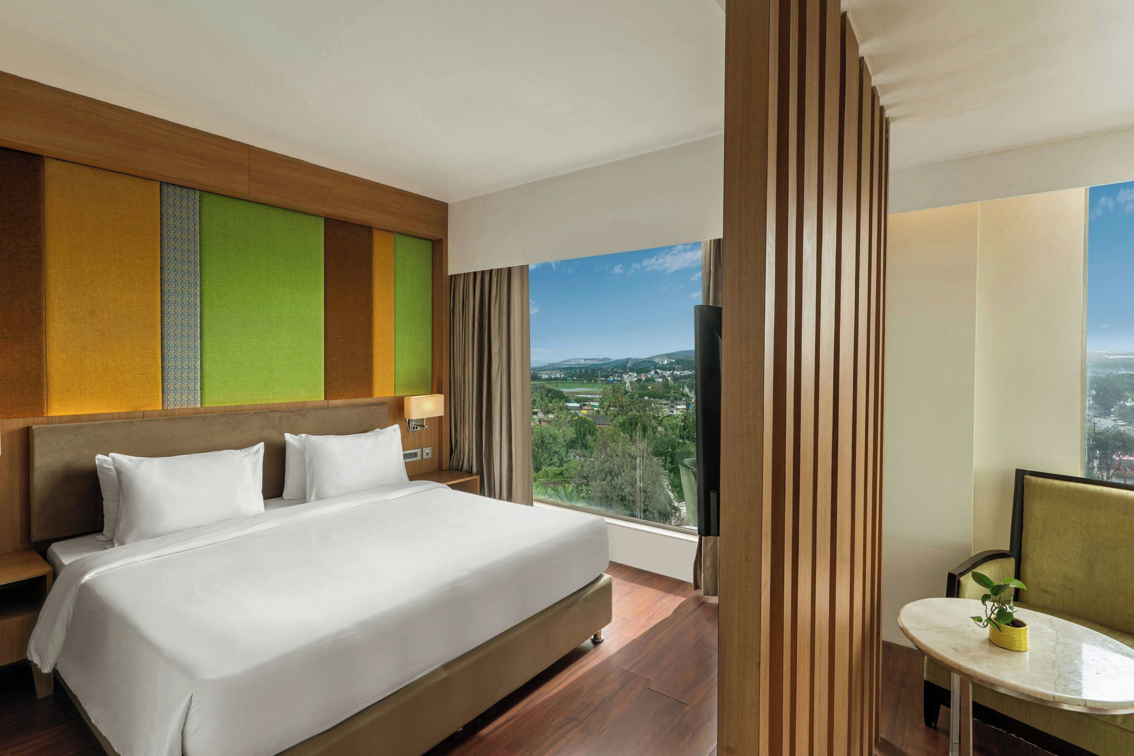 A hotel room with a large bed, a view of the city, and a small table with a plant.