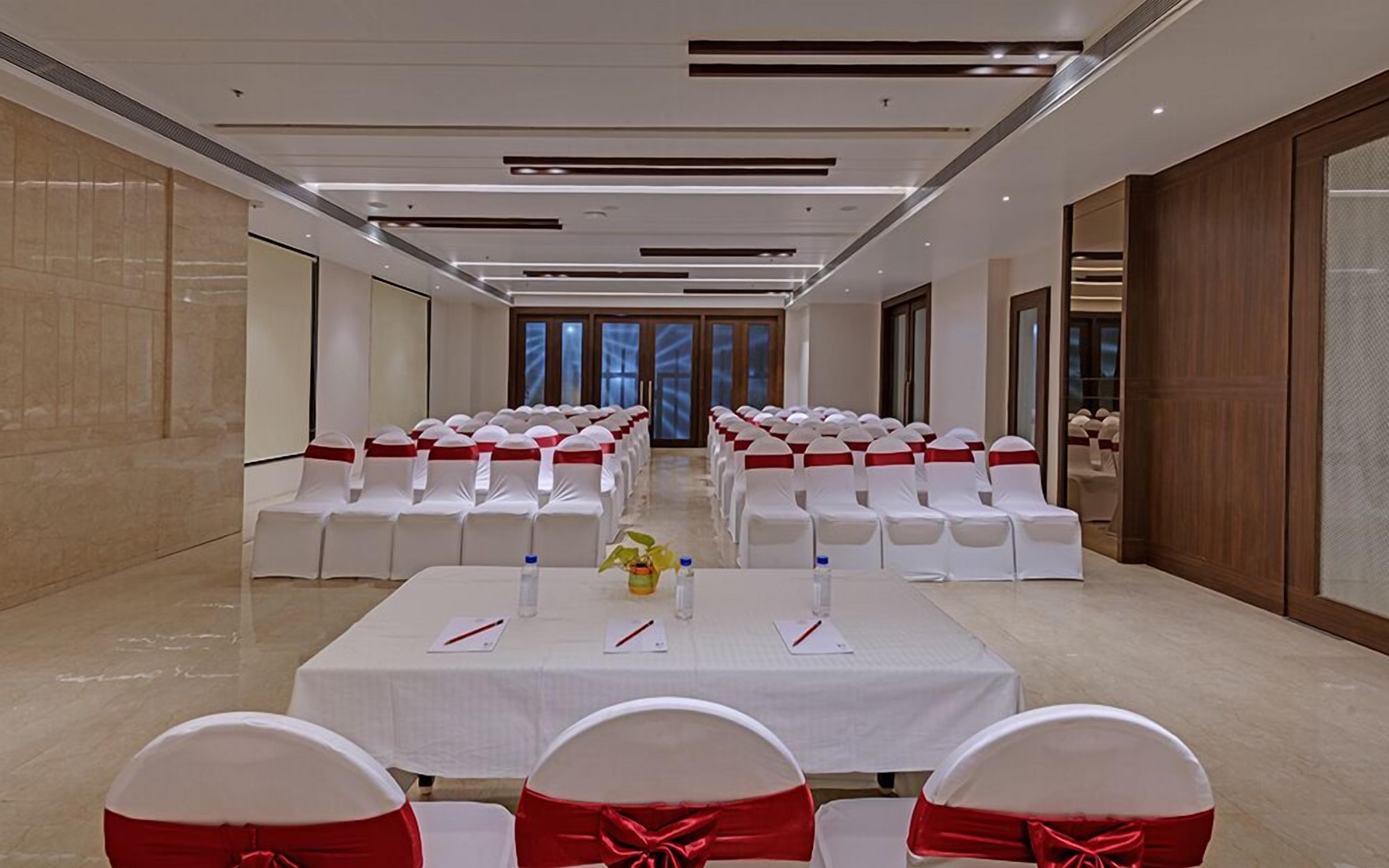 Un salón de conferencias con filas de sillas blancas adornadas con cintas rojas, una larga mesa con tarjetas de nombres, botellas de agua y una planta.
