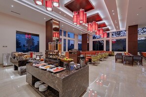 A spacious hotel dining area with a buffet setup, large windows, and modern red pendant lighting.