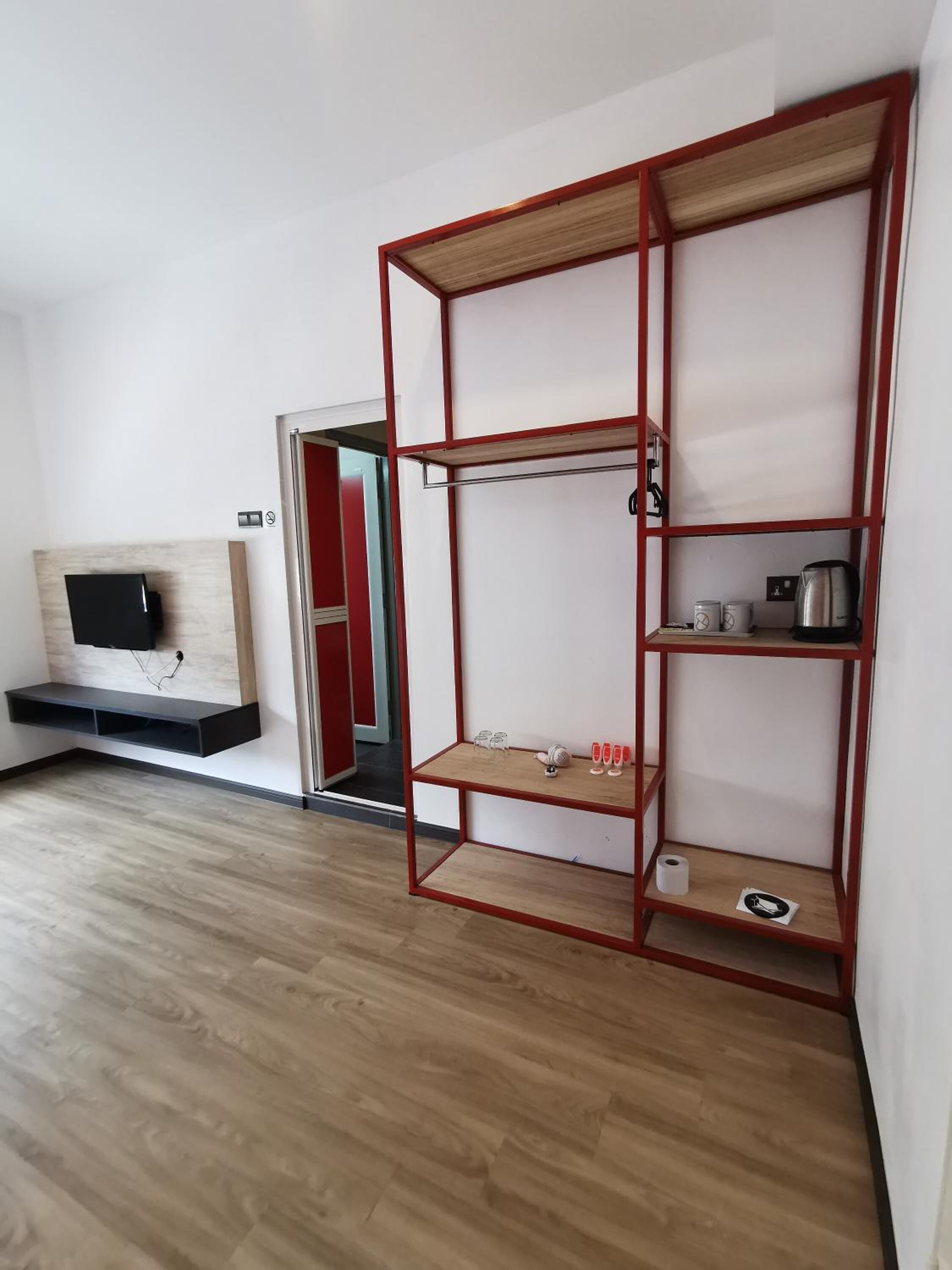 Una habitación con piso de madera, un televisor montado en la pared y una estantería metálica roja con varios objetos encima.