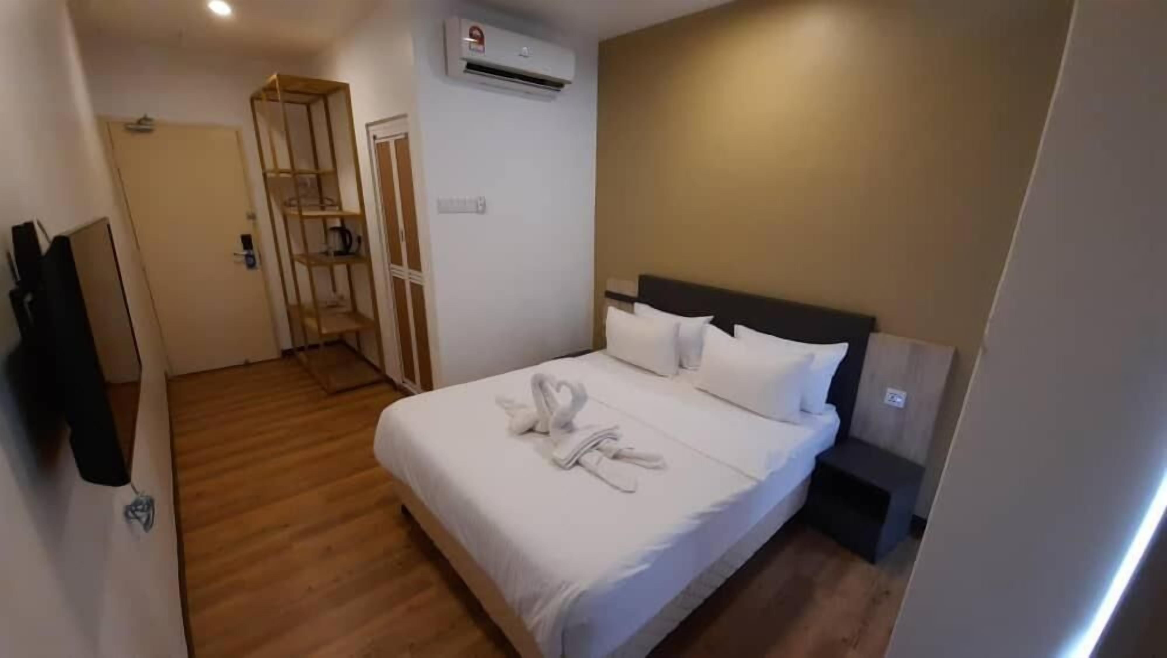 A hotel room with a wooden floor, a bed with white bedding and a heart-shaped towel arrangement, a bedside table, and a TV mounted on the wall.