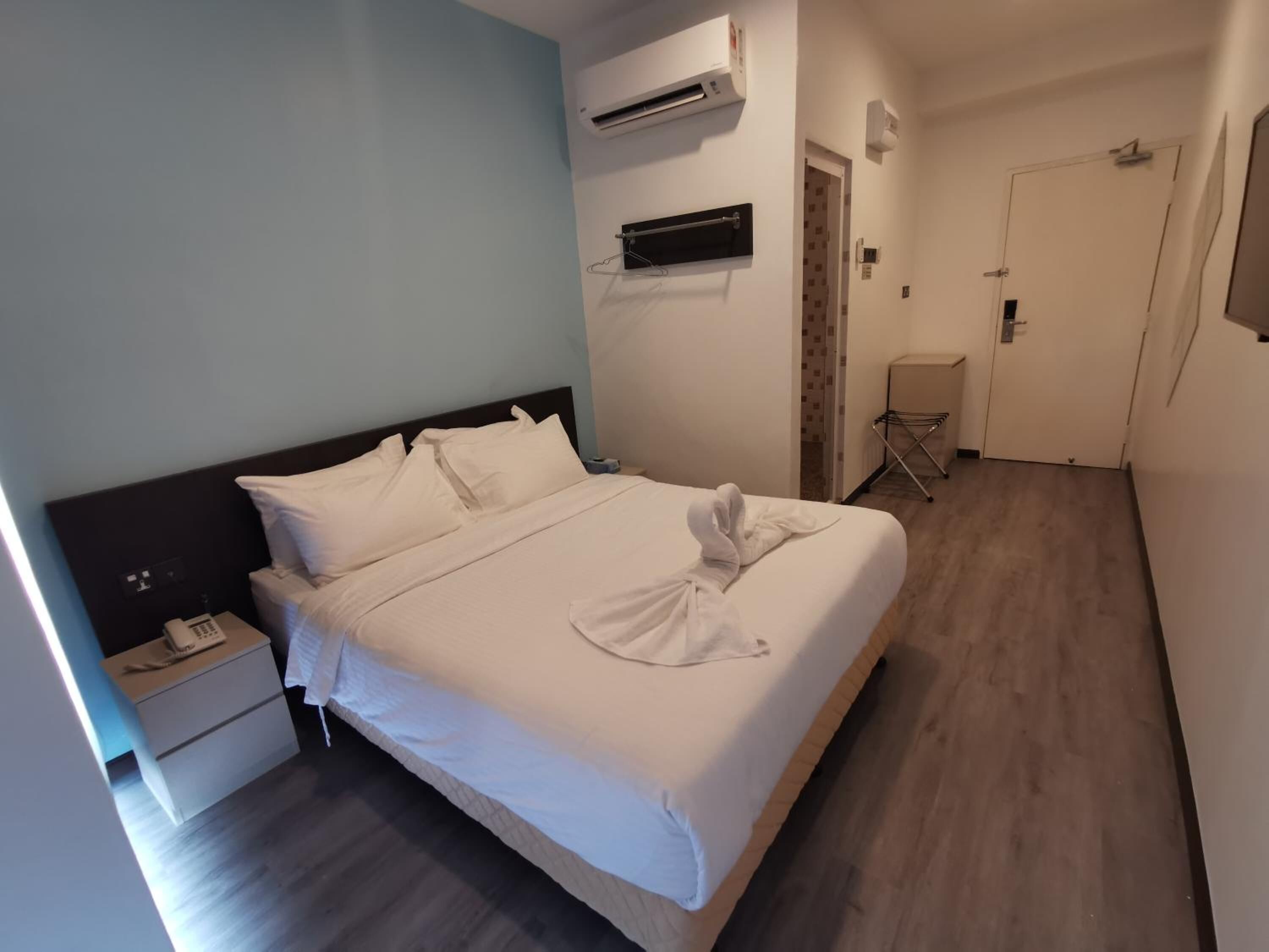 A hotel room with a bed, a nightstand, an air conditioning unit, a door, and a small seating area.