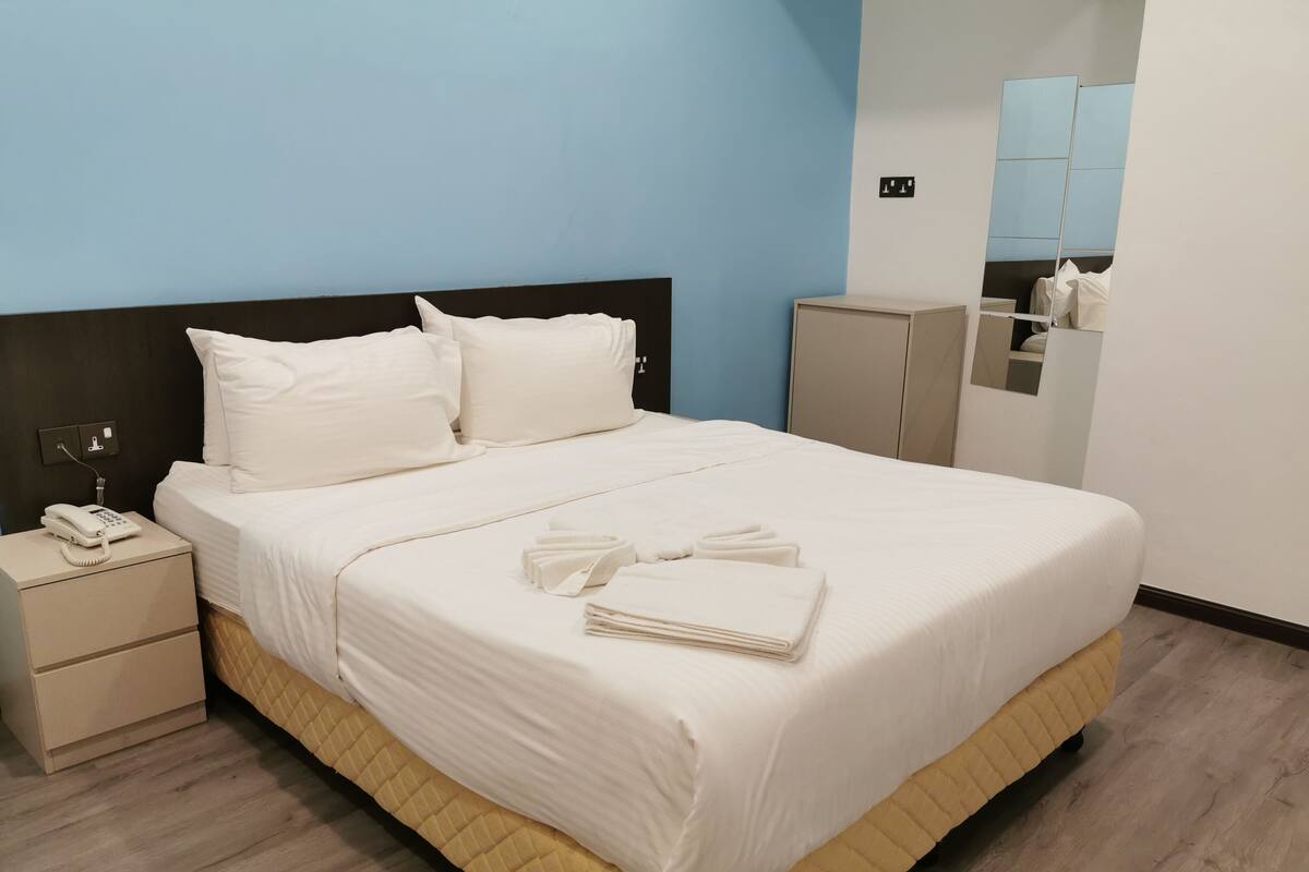 A neatly made bed with white linens, a bedside phone, and folded towels on a wooden floor.