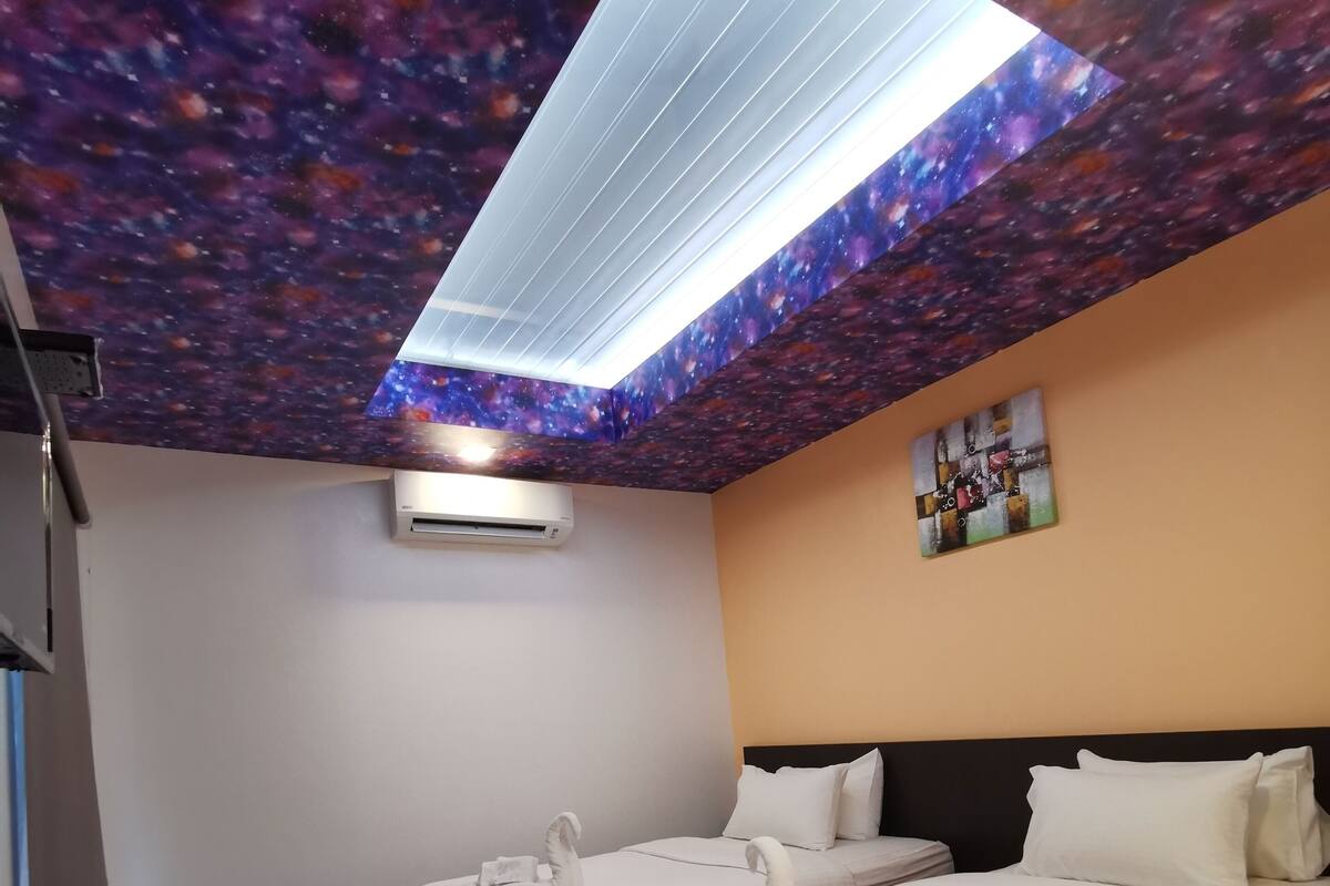 A hotel room with two beds, a skylight with a galaxy-themed ceiling, an air conditioning unit, a small table with a trash bin, and a painting on the wall.