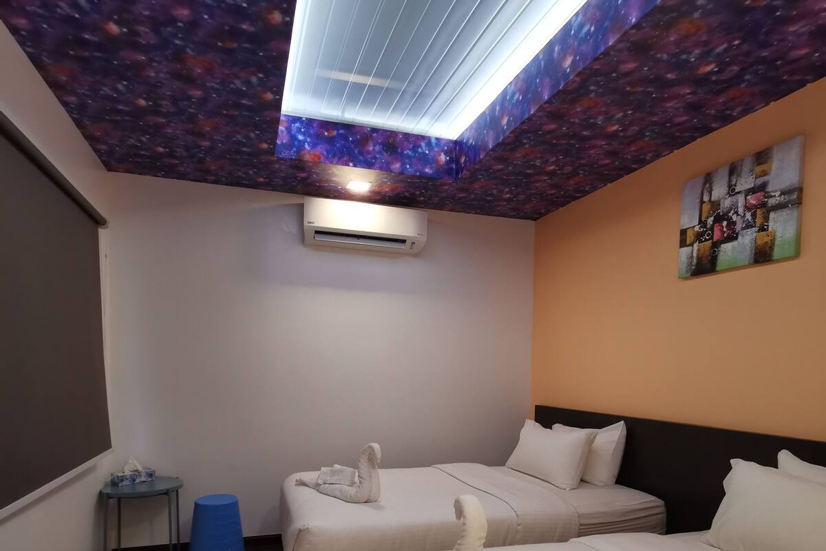 A hotel room with two beds, a ceiling with a galaxy-themed design, an air conditioning unit, a small table with a blue stool, and a wall-mounted artwork.