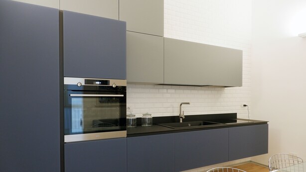 A modern kitchen with grey cabinets, a black countertop, a stainless steel oven, and a white tiled backsplash. - Magdahause (Taranto)