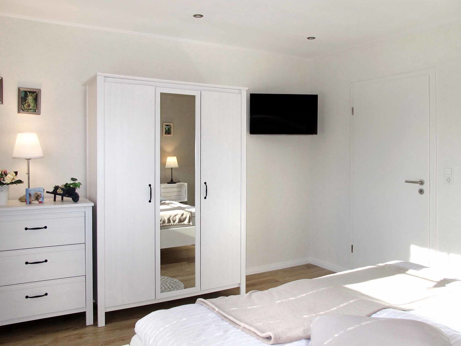 Ein Schlafzimmer mit Bett, Kleiderschrank mit Spiegel, Nachttisch mit Lampe und einem an der Wand befestigten Fernseher.