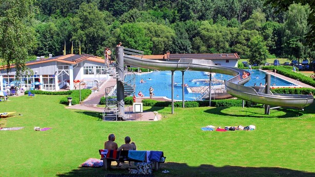 Ein Poolbereich mit Wasserrutsche, umgeben von einer Rasenfläche und einem Gebäude mit rotem Dach.
