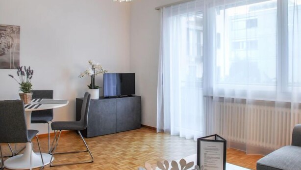 A modern living room with a wooden floor, a grey sofa, a dining table with chairs, a grey cabinet with a TV, and a large window with sheer curtains. - Junior Suite by Interhome (Ascona)