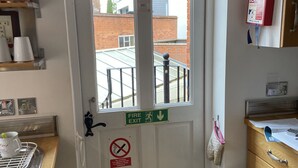 A kitchen with a white door, a fire exit sign, a no-smoking sign, a trash bin, and a window with a view of buildings. - 19 Station Road (Henley-on-Thames)