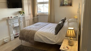 A bedroom with a bed, bedside table with a lamp, a TV on the wall, and a window with curtains. - 19 Station Road (Henley-on-Thames)