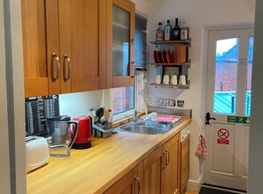 A kitchen with wooden cabinets, a red kettle, a sink, and a fire exit sign. - 19 Station Road (Henley-on-Thames)