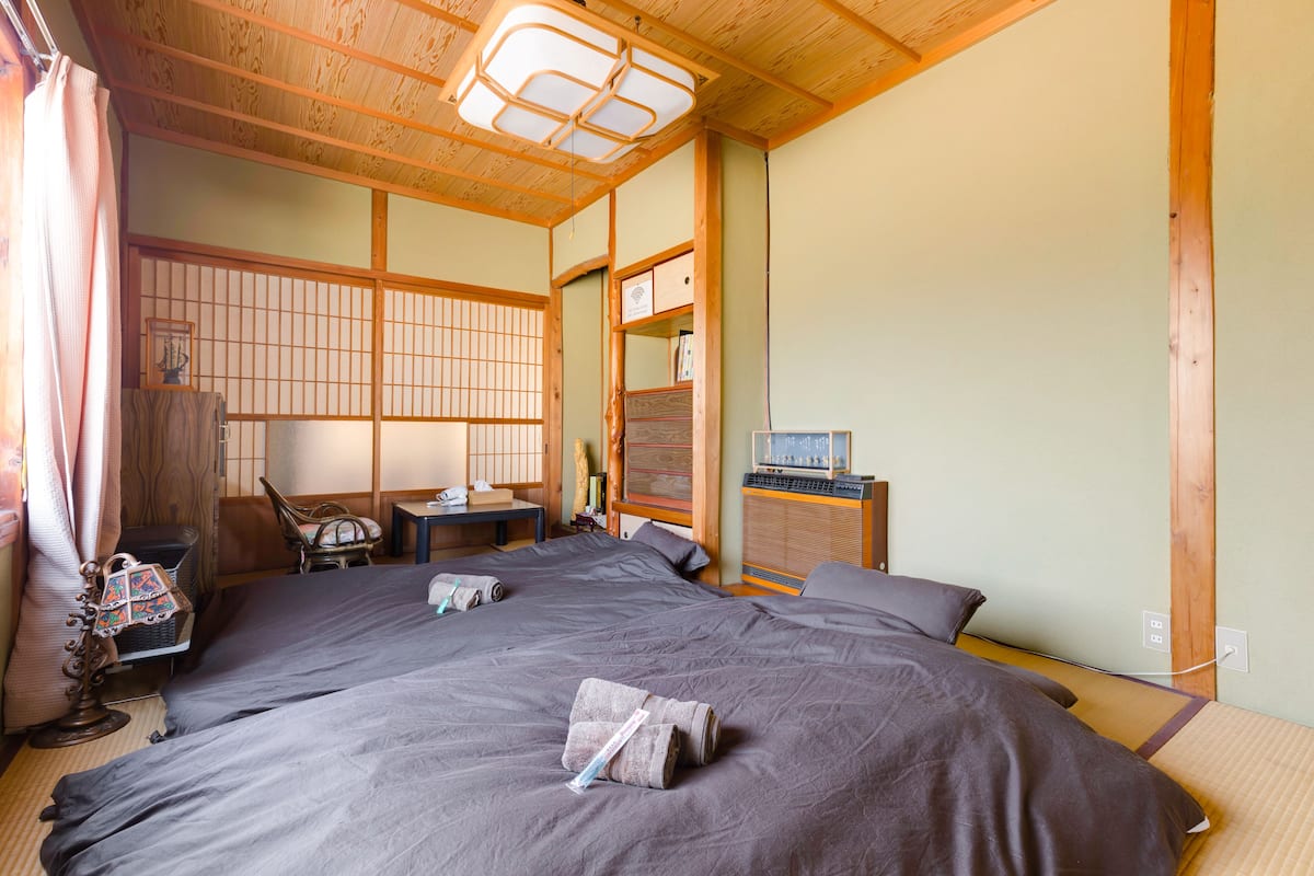 Una habitación tradicional japonesa con suelo de tatami, una cama grande con ropa de cama oscura, un escritorio de madera y un celosía japonesa.