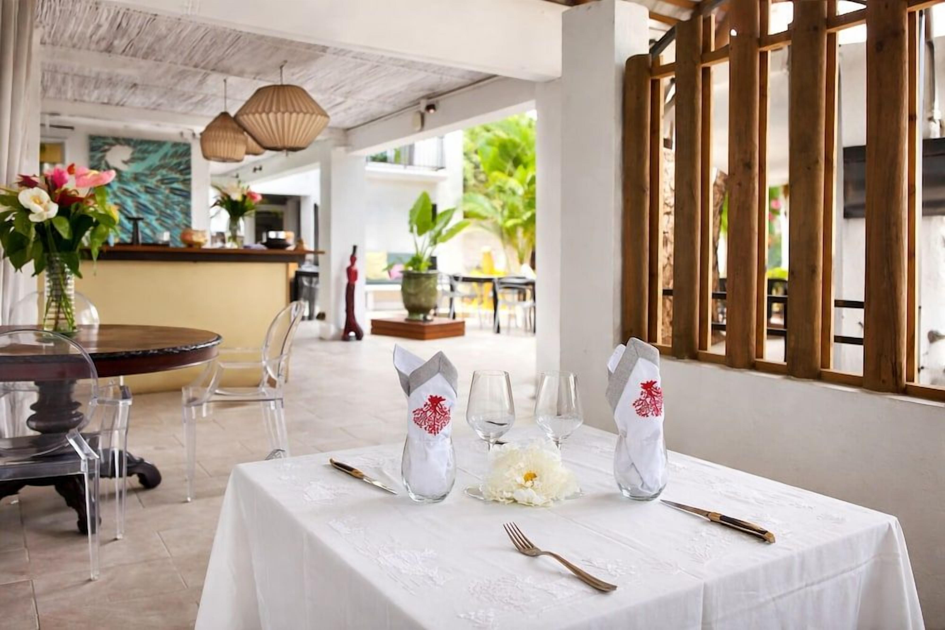 Un área de comedor al aire libre con una mesa redonda preparada para dos personas, sillas transparentes y un mantel blanco.