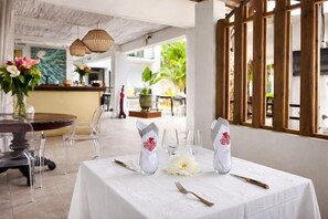 An outdoor dining area with a round table set for two, clear chairs, and a white tablecloth. - Sous le Badamier (pointe aux canonniers)