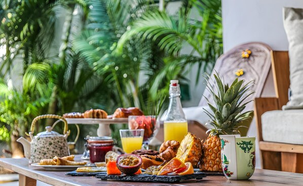 Una mesa de desayuno con frutas tropicales, pasteles y bebidas, frente a un fondo de vegetación exuberante.
