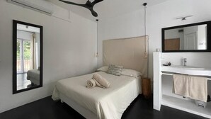 A bedroom with a bed, a ceiling fan, a large mirror, and a bathroom with a sink and towel. - Sous le Badamier (pointe aux canonniers)