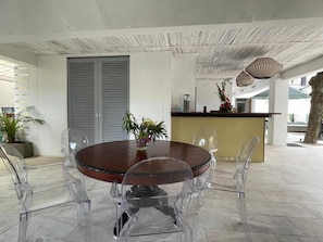 A modern dining area with a round wooden table surrounded by clear chairs, a kitchen area with a counter and decorative plants, and a ceiling with a unique textured design. - Sous le Badamier (pointe aux canonniers)