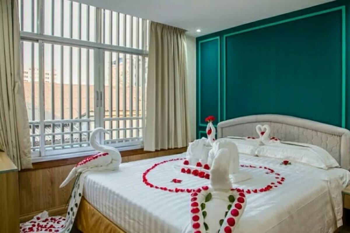 A hotel room with a bed adorned with swan decorations and rose petals, a window with a view of buildings, and teal-colored walls.